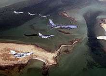 Photo of seven modern aircraft of differing designs flying in formation above a group of sandy islands