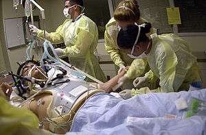 Health care providers attending to a person on a stretcher with a gunshot wound to the head, the patient is intubated, and a mechanical ventilator is visible in the background