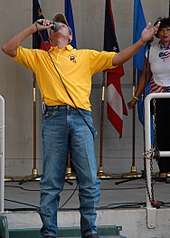 A man in a baseball cap, yellow T-shirt and blue jeans singing into a microphone with his arms spread wide