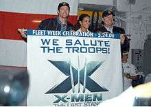 Hugh Jackman, Halle Berry and Kelsey Grammer hold a flag with the X-Men: The Last Stand logo and the inscription "We Salute Our Troops" in a ship's deck.