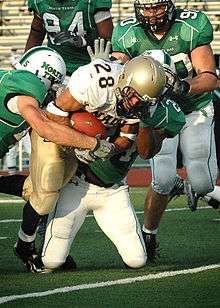 An American Football player in a gold and white uniform attempts to run past three defenders in green and white uniforms.