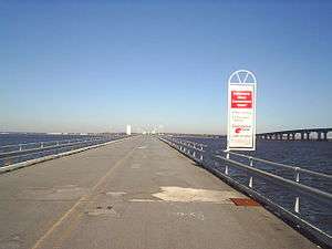 A two lane road on a low bridge over a large body of water with a higher span seen to the right