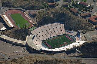 An aerial view of a stadium with blue end zones that appears build into the hills around it. There is a separate track field with seats above and to the left of the stadium.