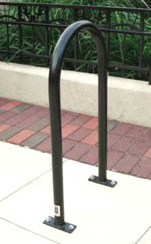 A tube of black-painted metal bent into a tall U shape and bolted at both ends to a concrete slab, in front of a brick pathway and iron railings