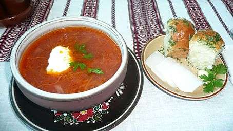 A bowl of red borscht served with two garlic pampushky and three slices of salo on a separate plate
