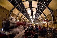 On the left is a red steam locomotive and three carriages, with steam drifting out above.  On the right passengers queue behind barriers and signs that say "Hogwarts Express" and "Platform 9¾".  Over both of these is a large cylindrical glass and steel roof.