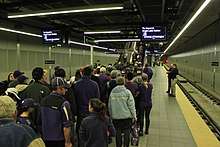 A crowd of people queue before an escalator between a set of empty train tracks