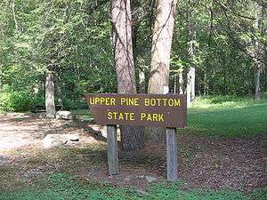 A brown wooden sign with Upper Pine Bottom State Park in yellow letters, with large tree trunks, rocks, and a picnic table in the background