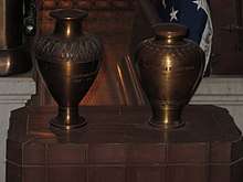 Two bronze urns sit on a table in front of two coffins. The urn on the left is slightly taller than the one on the right.