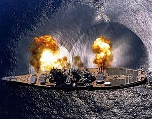 An overhead view of a battleship, showing the ship's teardrop shaped hull. Nine gun barrels on three turrets are pointed to the top of the image, with smoke and fire visible form all nine gun barrels and a concussive effect on the water below the guns. Visible from the overhead image are the 8 armored box launchers for the battleship's 32 Tomahawk Missiles.