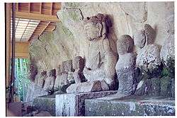 Photograph of a row of ca. ten seated stone statues in front of a rock. Three-quarter view. One of the statues is about twice as large as the others.