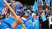 Demonstrators dressed in light blue, holding blue flags with white crescent. One in the foreground is wearing blue face paint.