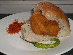 A plate of Vada Pav with seasoning of red chilli powder and a green chilli.