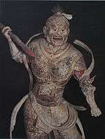 Frontal view of a fierce looking statue dressed in armour and wielding a stick-like object in his right hand.