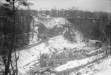 Construction across a bare valley; a bridge being built. The ground is covered in snow. In the background, an iron tressel carries cars over the valley.