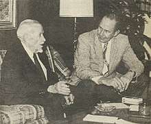 A black-and-white photograph of two men sitting by a low table talking to each other. The man on the left is much older, has white hair, and is wearing a dark suit with a white shirt and a dark tie. He sitting on a plaid couch and gesturing with his right hand as he speaks. The man on the left is younger, has dark hair, and is wearing a light jacket, dark pants, a white shirt and a patterned tie. He is sitting on a chair with his arms resting on his legs as he leans forward to listen to the other man.