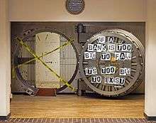 A large circular metal-lined opening set on a metal bed in a beige wall beneath an ornate silver wall clock reading 10:12. Inside is a space with bars on either side; entry is impaired by two crossed strips of yellow tape with "caution" written on it in English and Spanish. To its left is a heavy open door with metal workings behind a glass plate; pieces of paper with individual letters are affixed to it spelling out "If a bank is too big to fail, it is too big to exist"