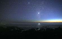 A photograph of the night sky taken from the seashore. A glimmer of sunlight is on the horizon. There are many stars visible. Venus is at the center, much brighter than any of the stars, and its light can be seen reflected in the ocean.