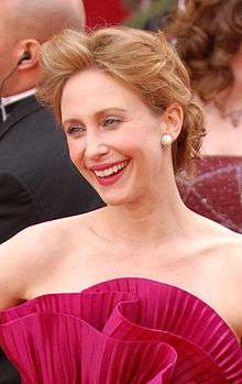 A light-skinned woman with red hair smiles while wearing a red dress, with other people in the background.