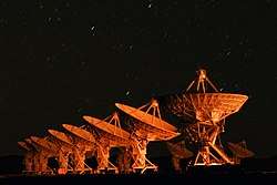 Orange light illuminates the array of dishes at night, with streaks of star-trails overhead.
