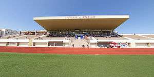 The stand of a football stadium, appearing to be made of concrete, in the daytime. The centre section of the stand is covered by a roof. On the front edge of the roof, the letters "VICTORIA STADIUM" can be seen.