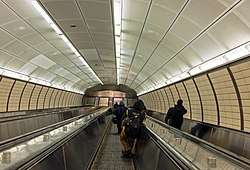 Looking down a bank of three escalators in an elliptical passage lit by fluorescent light strips above, generally white with some yellow paneling on the walls. There are several people on the escalator at varying distances from the camera
