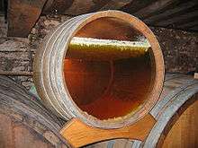 Photo showing a barrel with a glass bottom revealing the layer of yeast on the surface under which the yellow wine is ageing. This yeast is in the form of an irregular white layer – it forms small stalactites that sink a few millimetres into the wine which is already dark yellow wine.  The wall in the background is grey, probably coated in grey mould.
