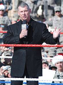 A man with short gray hair wearing a black suit jackets and black pants speaks into a microphone while standing in the middle of a wrestling ring, gesturing with his left hand. Behind him, a crowd of men wearing army camouflage uniform watches.