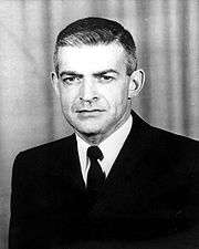 Head and shoulders of a white man with short hair neatly combed, wearing a dark suit coat and tie.