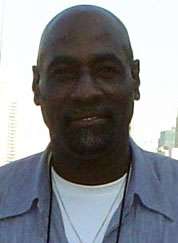 A black skinned man with a bald head and black goatee beard. He is wearing a light blue shirt with the top buttons undone, showing a white top underneath.