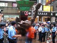 huge bronze sculpture being taken away by removal men dressed in orange, surrounded by police officers in blue shirts and caps or berets and overlooked by crowds