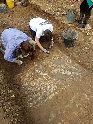 two people on their knees, in a trench, brushing dirt from the mosaic
