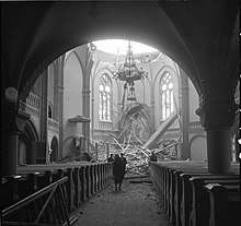 The dome of the Vyborg Cathedral has collapsed after Soviet bombing. Four people stand in the nave and look at the rubble, highlighted by sunlight shining through the damaged dome.