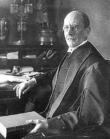 A middle aged white man in judicial robes holds a book with his left hand, rests his right on a table, and looks towards the camera.