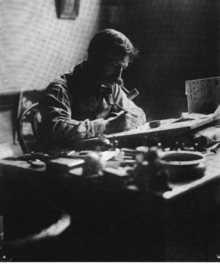 Black-and-white photograph of a man with a large mustache and a pipe in his mouth, at work, seated behind a cluttered desk on which a drawing board is propped at a slight angle.