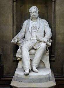 A white statue of a seated middle-aged man in mid-19th Century clothing, holding rolled plans in his left hand
