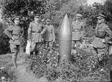 A group photograph of several men in uniform who are standing around an unexploded shell that is standing pointed-end up from the ground.