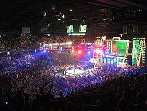 A sold out crowd watching the event, with a professional wrestling ring in the center of the picture. A giant screen in the shape of a briefcase is at the top right.