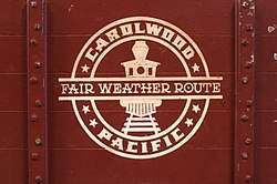 A circular, white-colored logo consisting of a forward-facing steam locomotive in the center, the text "Carolwood Pacific" around the edge, and the text "Fair Weather Route" across the middle. The logo is painted on the side of a miniature, reddish-brown-colored freight car.