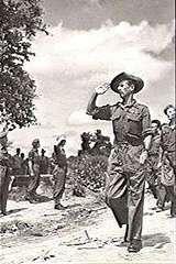 A marching soldier, in full military uniform and slouch hat, leading several other soldiers. He is saluting a stationary group to his right.