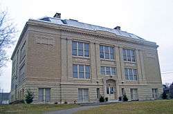A large square beige-colored three-story brick building with pilasters on the front and the words "Washington School" engraved below the roof. Between the windows on the second and third stories are the words "science" "language" and "history".
