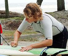 Photo of woman bent over surfboard rubbing bar of solid wax against the board with palm trees and ocean in background