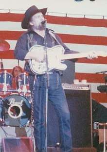 A man wearing a black cowboy hat, black shirt, black vest and blue jeans, playing a guitar and singing into a microphone