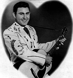 A dark-haired man wearing a suit with musical notes printed on it, playing a guitar