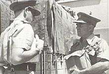 Two men in military uniform conversing next to a wooden structure. The man on the left is slightly side on with his back to the camera, while the man on the right is slightly shorter and holding a bag.