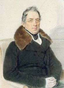 A man with thin grey hair sits in a chair, hands folded on his lap. He is wearing a grey coat with a brown fur collar.