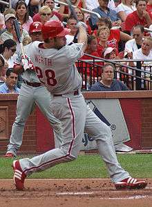 A right-handed man in a gray baseball uniform with red trim and a red batting helmet swings at a baseball with a baseball bat. His jersey reads "Werth" and "28" on the back in red letters.