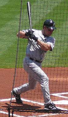 A man in a gray baseball uniform and black batting helmet stands in the batter's box twisting from the waist while holding a baseball bat