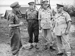 Four men in uniforms wearing peaked caps stand around talking. In the background are some tents and scaffolding.