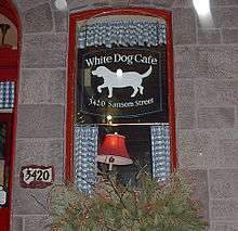 A photograph of a window with a black sign in it with white letters reading "White Dog Cafe 3420 Sansom Street" and a silhouette of a white dog facing left